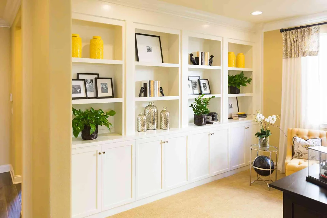 White built-in shelves with yellow vases, framed photos, books, green plants, and decorative objects in a bright, modern living room with beige walls and a window with patterned curtains.