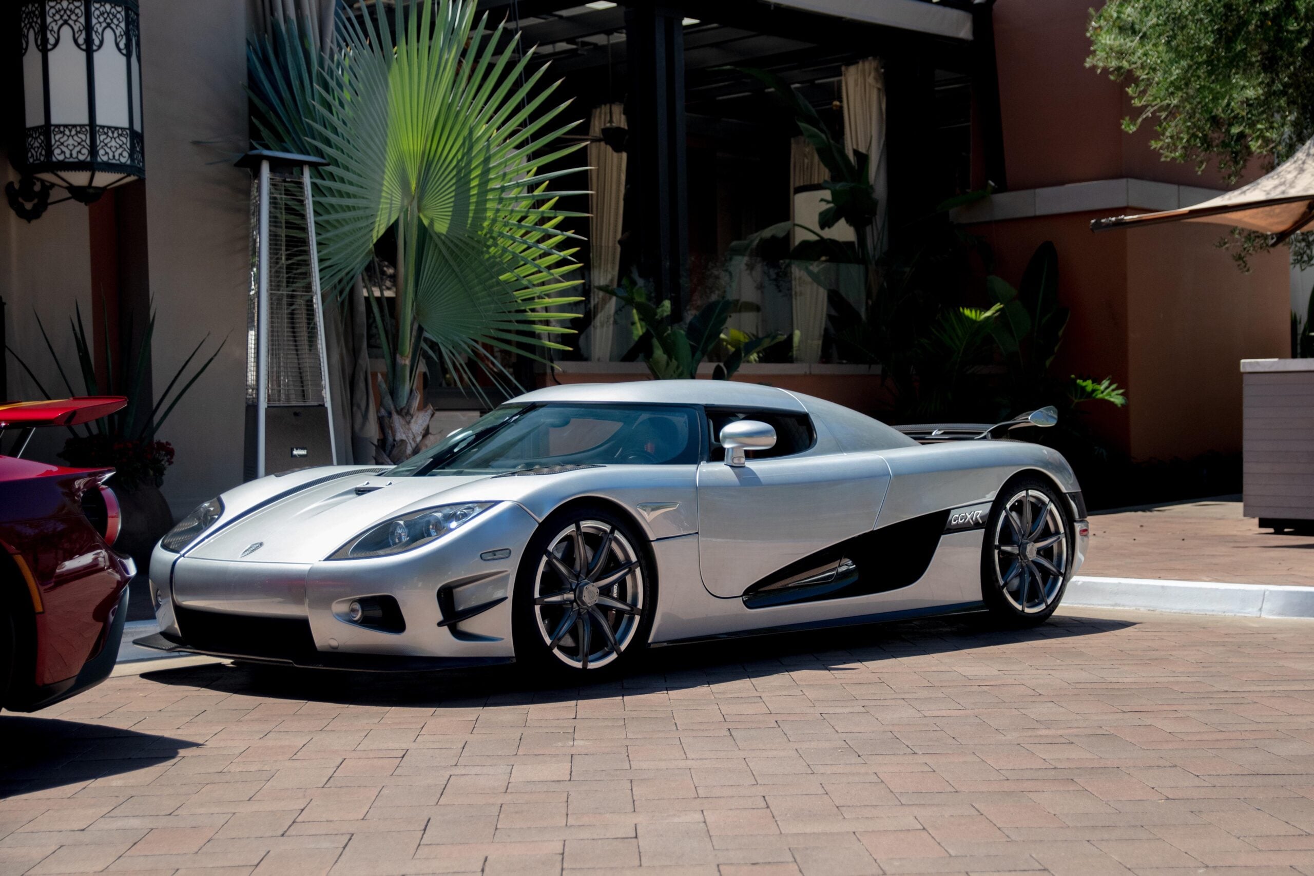 A silver sports car with black accents parked on a brick driveway in front of a building with large plants and windows.
