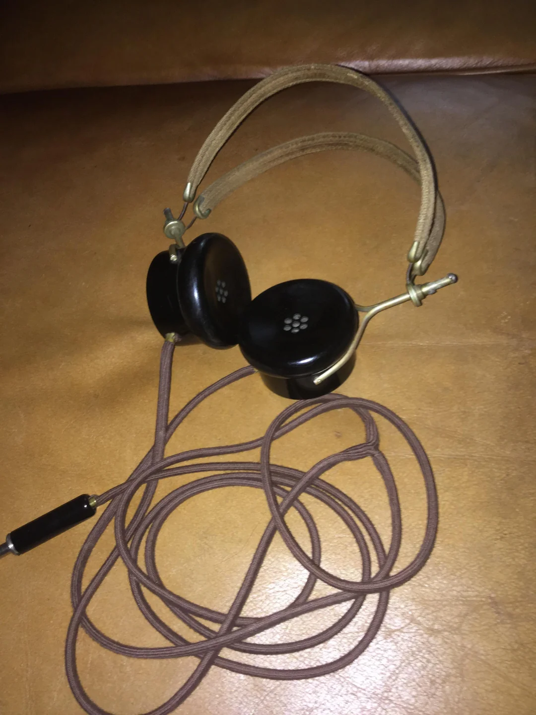 A pair of vintage over-ear headphones with a brown fabric-covered headband and cord, resting on a tan leather surface. The earpieces are black with small circular vents.