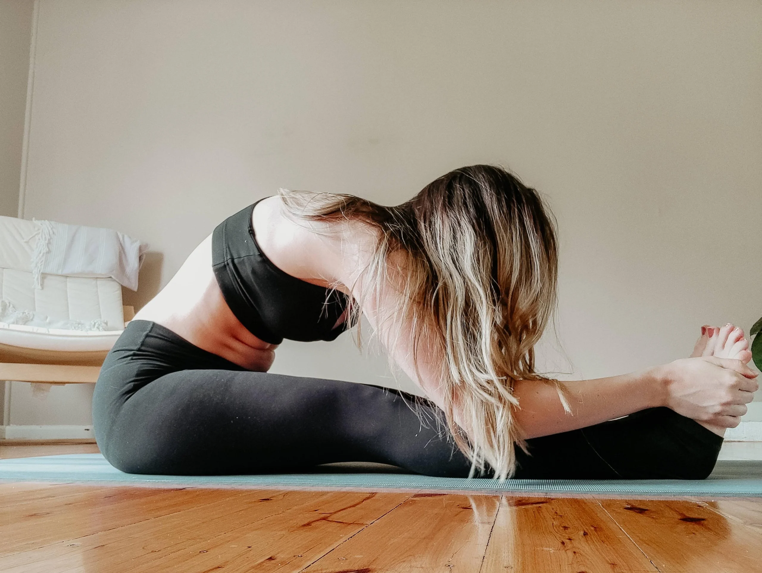 A woman wearing black athletic wear sits on a yoga mat indoors, stretching forward to touch her toes with both hands, her head down and hair falling forward.