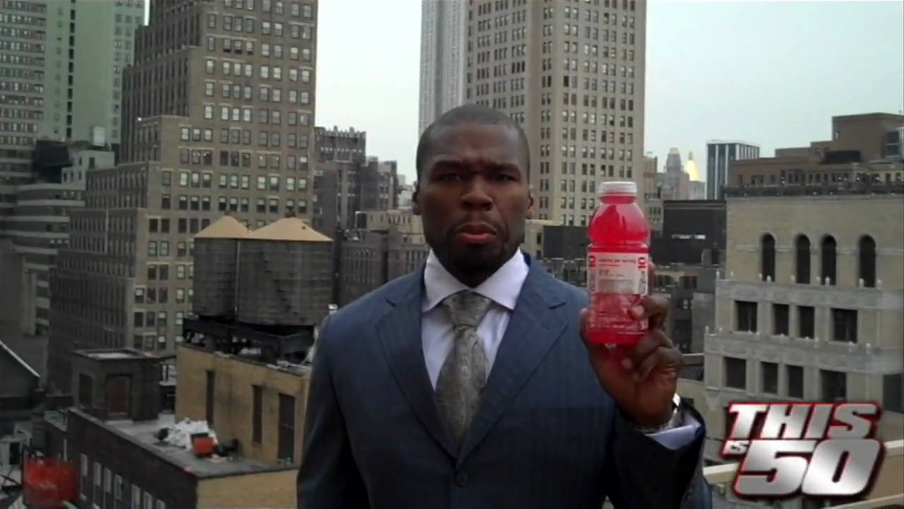 A man in a suit stands on a city rooftop holding up a red beverage bottle, with tall buildings in the background. The logo "THISis50" appears in the lower right corner of the image.