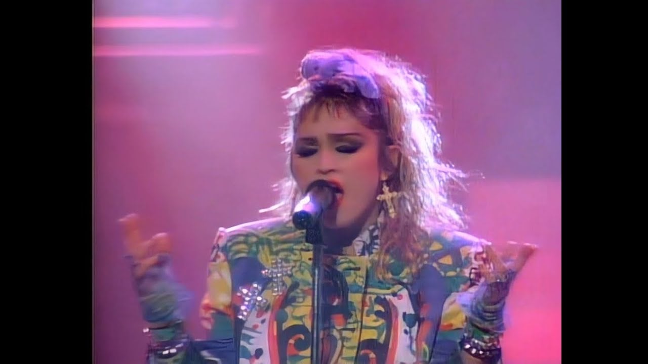 A singer with teased blonde hair, bold makeup, and colorful 1980s clothing performs passionately into a microphone on a pink-lit stage.