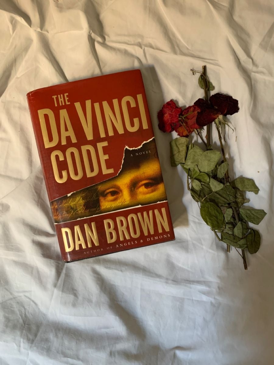 A copy of "The Da Vinci Code" by Dan Brown rests on a white bedsheet beside a bunch of dried red roses and green leaves.