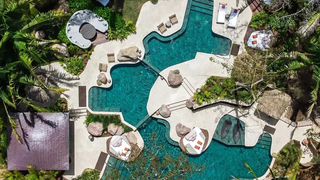 Aerial view of a tropical resort pool with curved shapes, surrounded by lounge chairs, umbrellas, greenery, and palm trees. Several small bridges and rock features cross over and border the water.