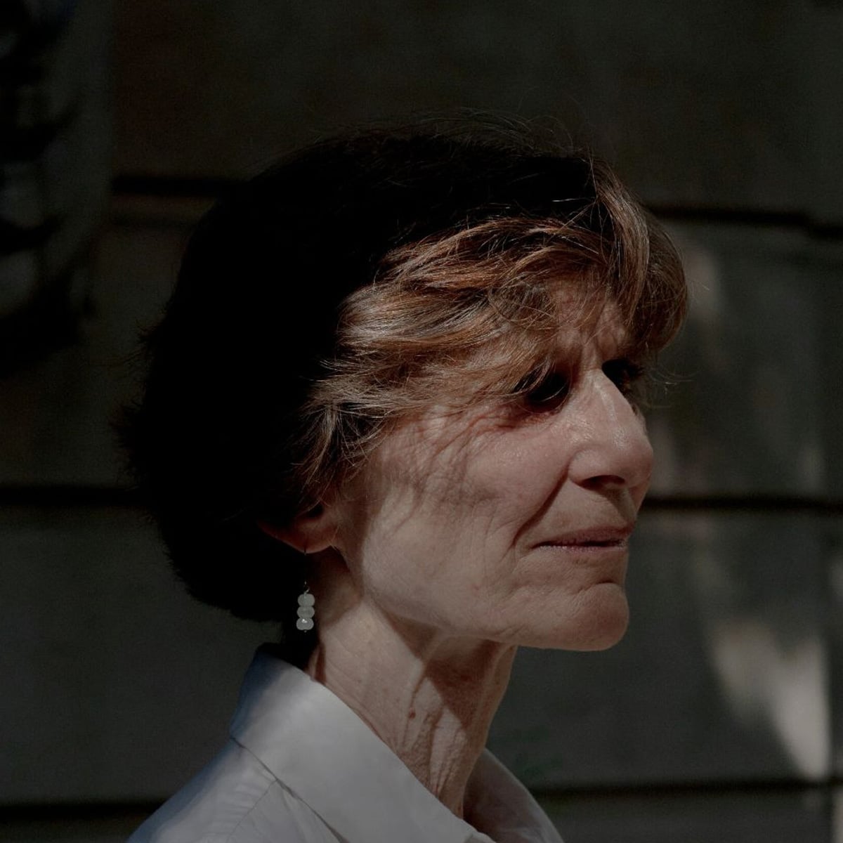 A woman with short, dark hair and light streaks stands in profile, wearing a white collared shirt and pearl drop earrings. She is lit by soft, natural light with shadows falling across her face and a blurred background.