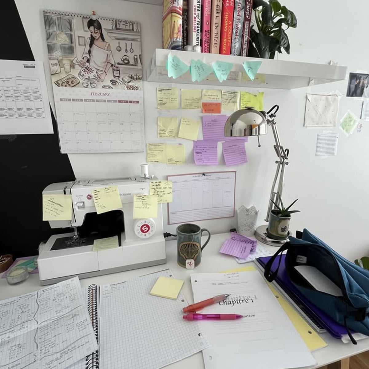 A cluttered desk with a sewing machine, notebooks, pens, a mug, sticky notes on the wall, a lamp, books on a shelf, and a calendar featuring a woman cooking. There is also a plant and an open pencil case.