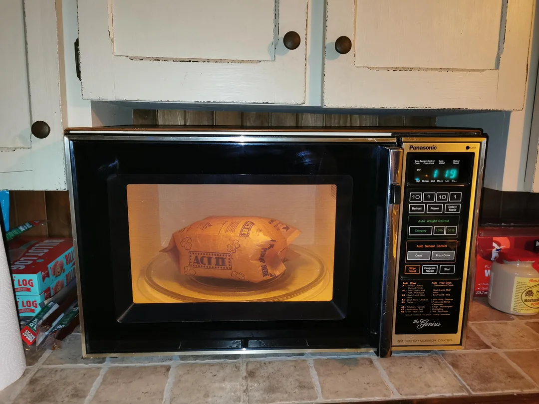 A bag of popcorn is being microwaved inside a vintage Panasonic microwave oven on a kitchen counter with cabinets and other items visible in the background.