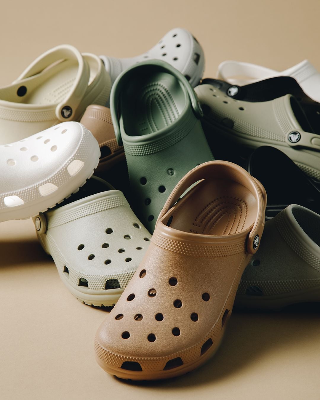 A pile of Crocs-style clogs in various neutral colors, including tan, green, white, and beige, arranged on a beige background.