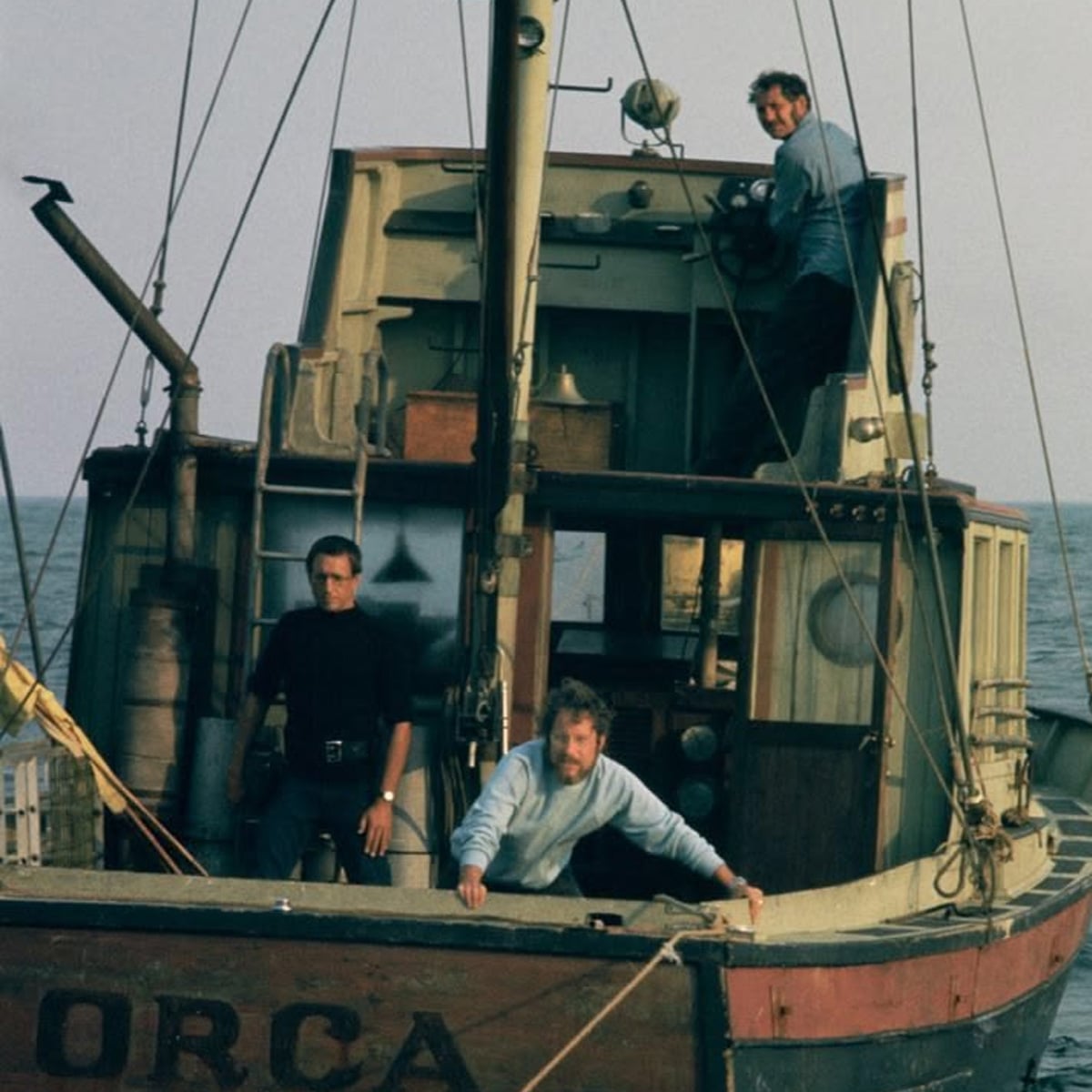 Three men are standing on an old fishing boat named “Orca” at sea. One man is steering at the top, while the other two stand on the lower deck, looking forward with serious expressions.