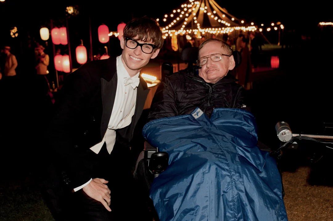 A smiling man in a tuxedo poses next to a man in a wheelchair who is wrapped in a blue blanket, with string lights and lanterns illuminating an outdoor nighttime setting in the background.