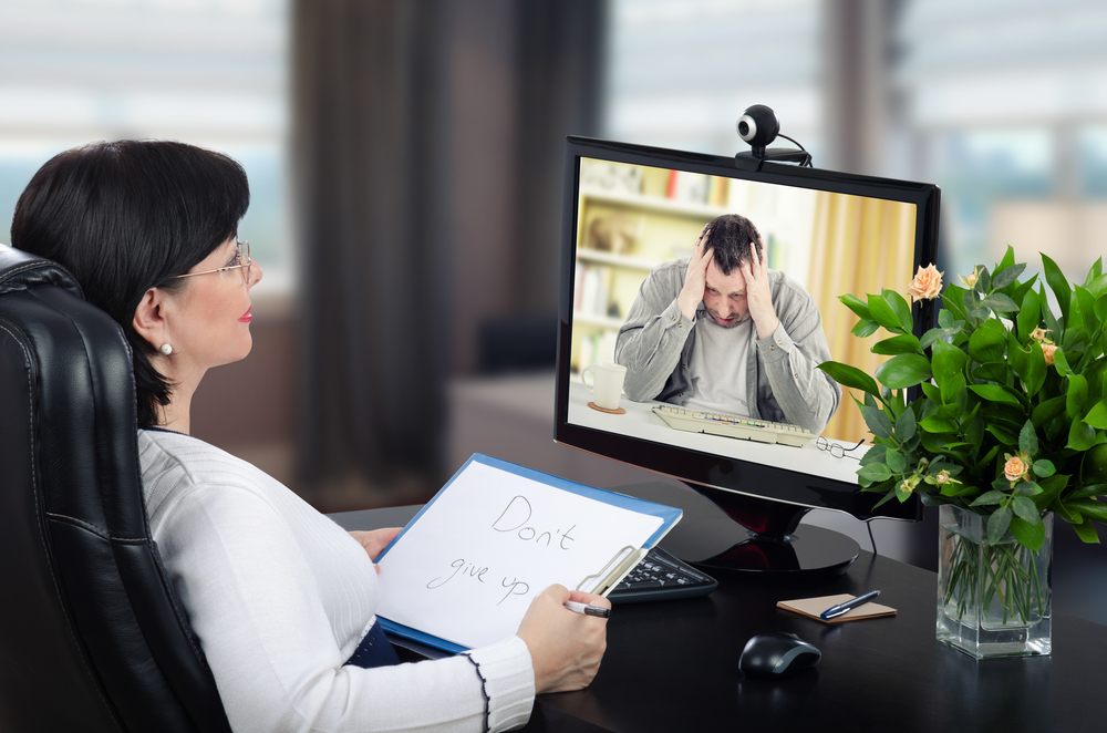 A woman sits at a desk holding a sign that says "Don't give up" during a video call with a distressed man, who is holding his head in his hands. A flower pot and computer mouse are on the desk.