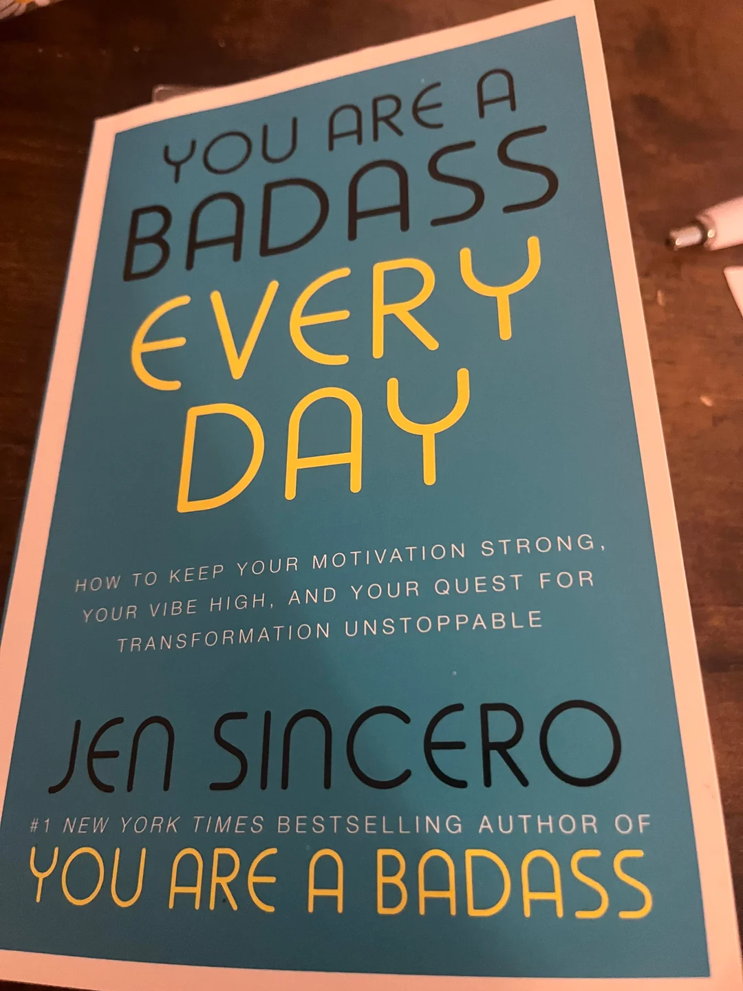 A teal book titled "You Are a Badass Every Day" by Jen Sincero, with yellow and black text emphasizing the words "Badass" and "Every Day," lies on a brown surface near a pen.