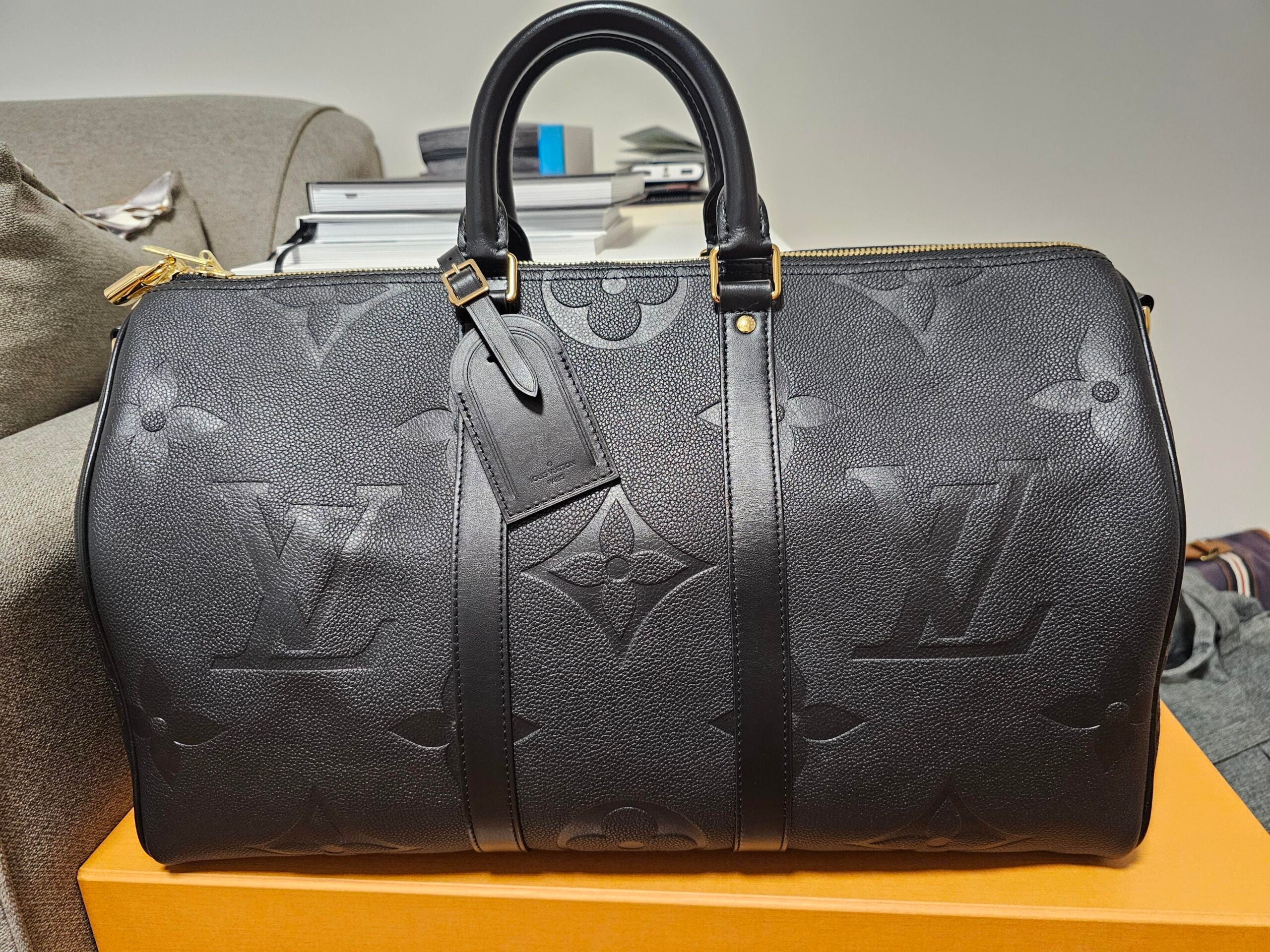 A black Louis Vuitton duffle bag with embossed LV monogram sits on an orange box, with a stack of books and a gray couch in the background.