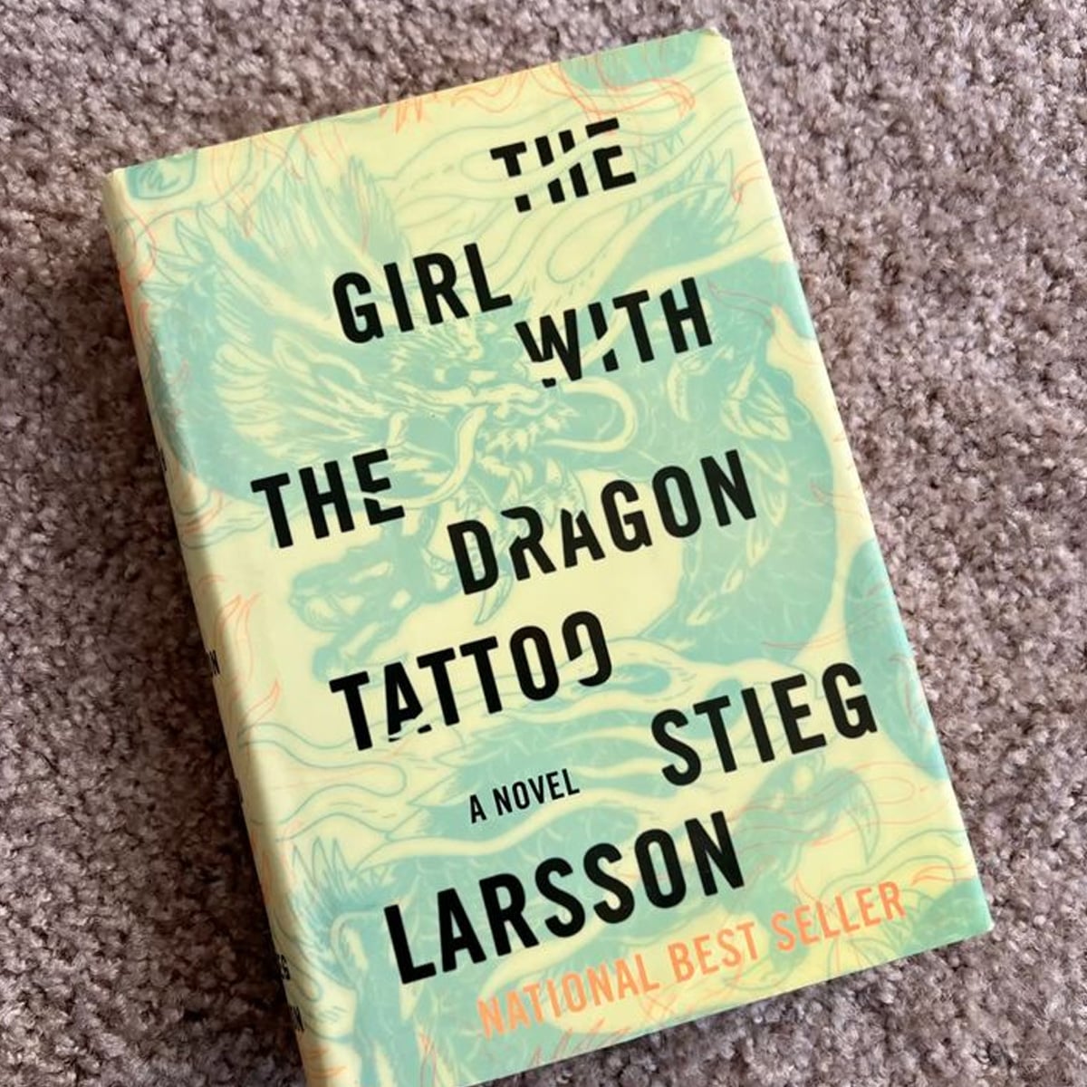 A hardcover copy of "The Girl with the Dragon Tattoo" by Stieg Larsson rests on a carpet. The book cover features bold black text and a pale green and yellow background with a faint dragon illustration.