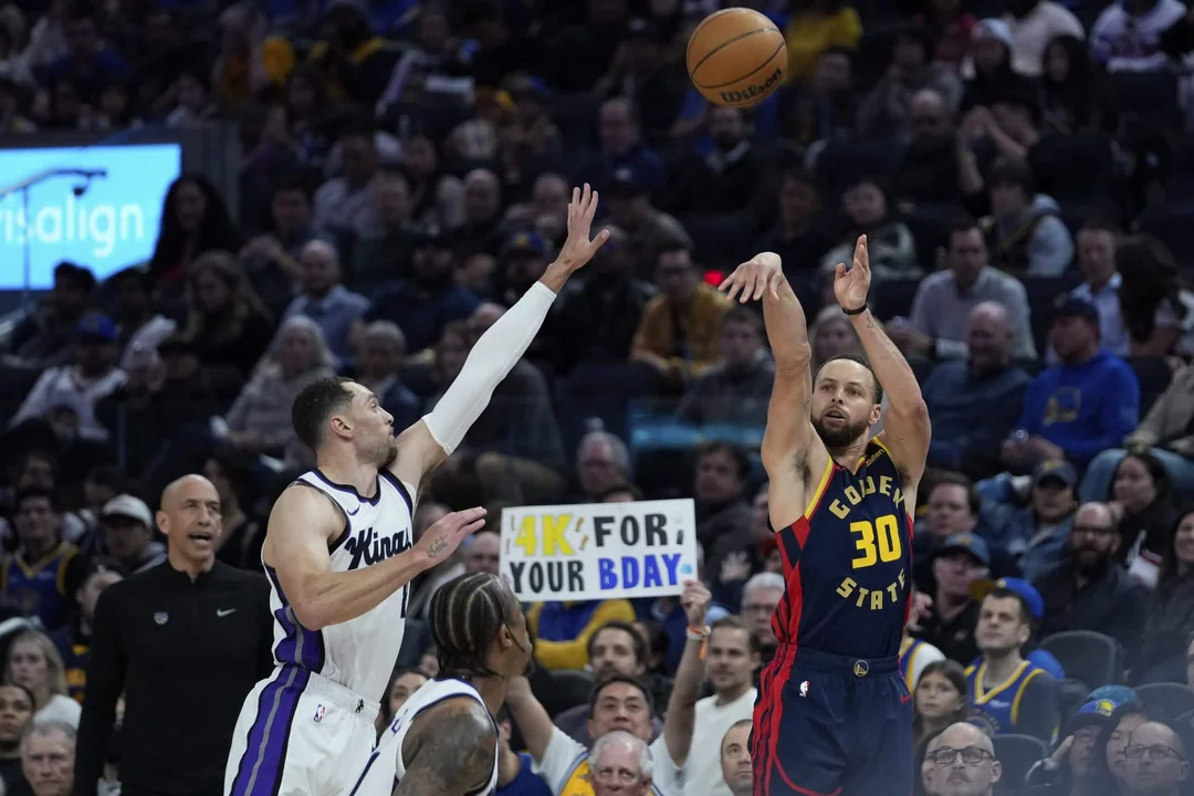 A Golden State Warriors player wearing jersey number 30 shoots a basketball over a Sacramento Kings defender during a game, with a cheering crowd and a colorful birthday sign in the background.