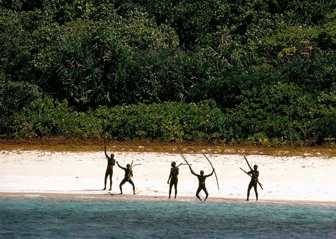 Five people stand on a sandy beach near dense green forest, holding long sticks and raising their arms, facing the camera. The scene is viewed from the water, with the shoreline and foliage in the background.