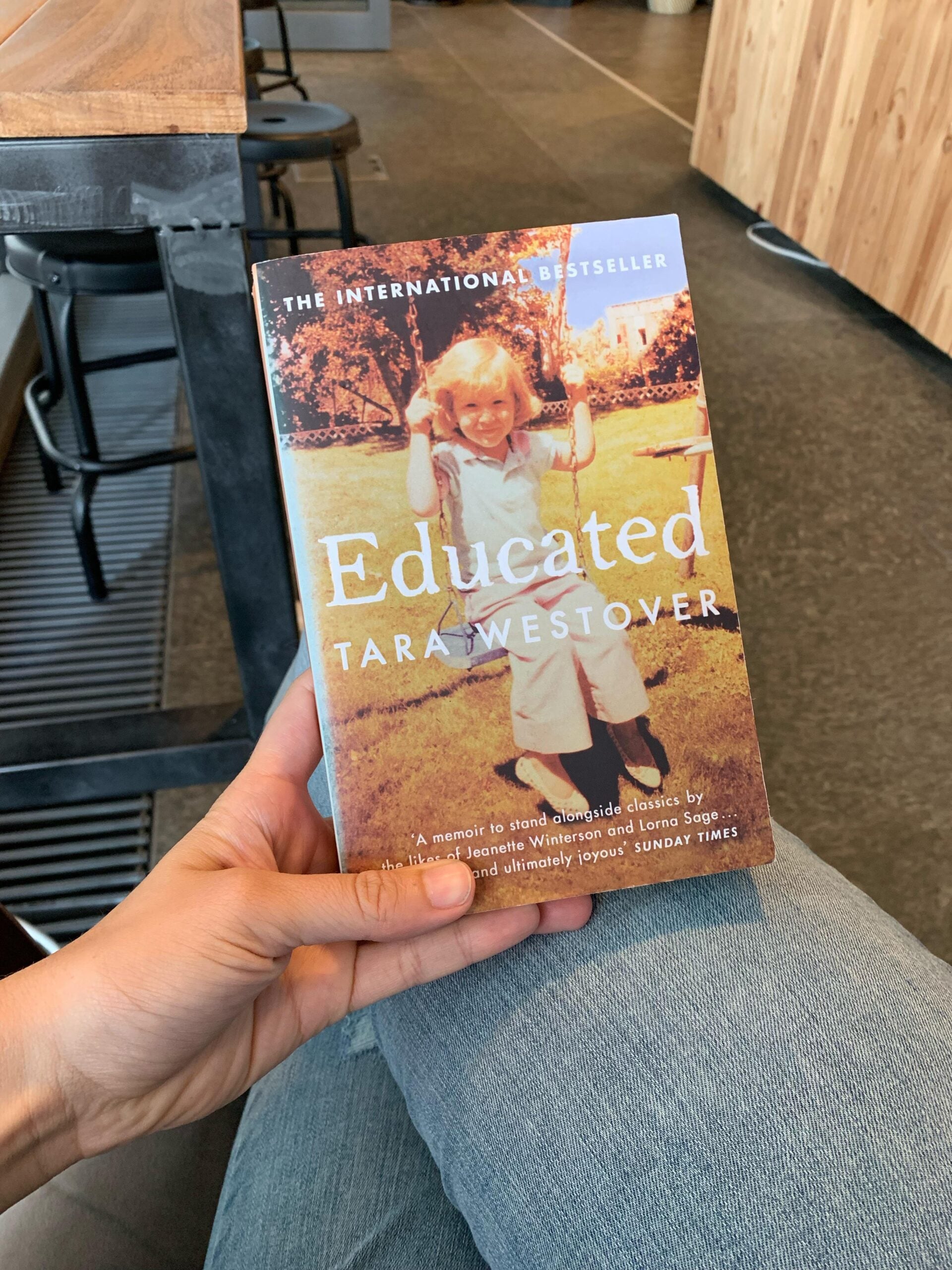 A person holds the book "Educated" by Tara Westover while sitting cross-legged in a modern café with wooden and metal furniture. The book cover features a young girl swinging outdoors.