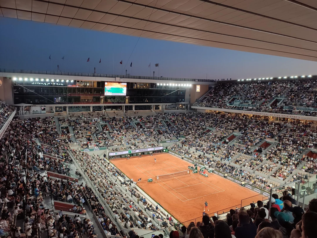 A large crowd watches a tennis match on a clay court in a modern outdoor stadium at dusk, with bright lights illuminating the scene and a scoreboard visible in the background.