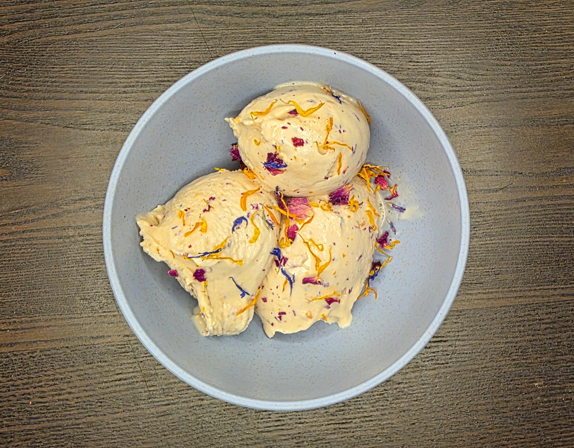 A bowl with three scoops of light-colored ice cream, garnished with colorful dried flower petals, sits on a wooden surface.