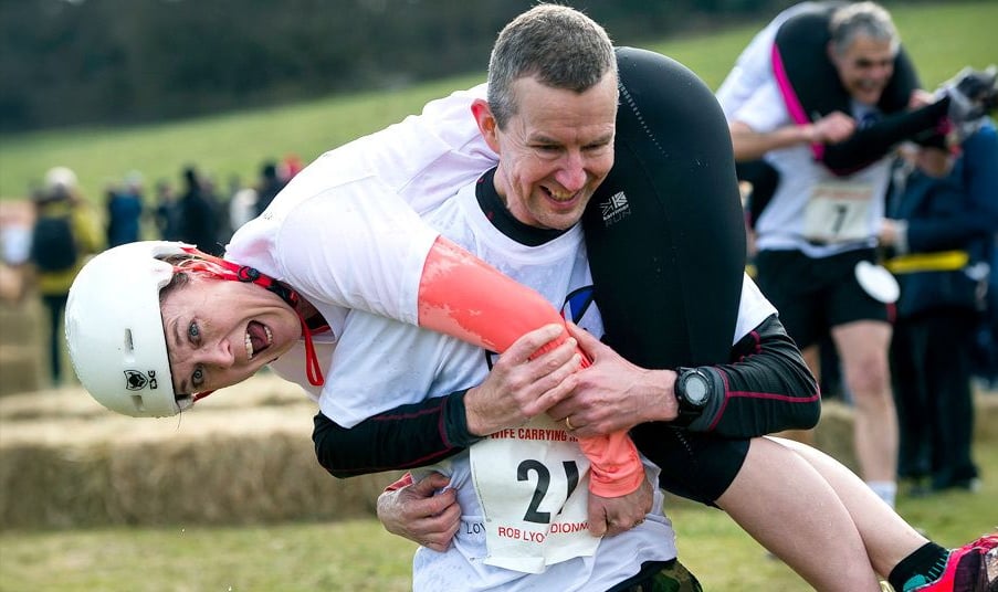 A man participates in a race by carrying a woman over his shoulders. Both wear athletic gear, the woman also wears a helmet. They are outdoors with other competitors and hay bales visible in the background.