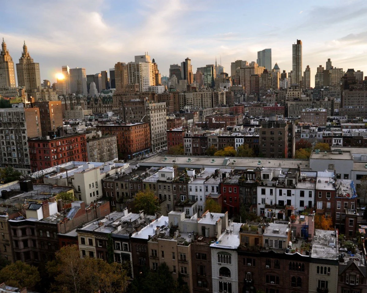 A cityscape showing rows of brownstone buildings in the foreground with high-rise skyscrapers in the background under a partly cloudy sky at sunset.