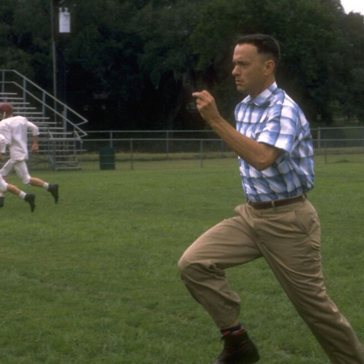 A man in a blue plaid shirt and khaki pants is running across a grassy field, with bleachers and another person running in the background. Trees surround the area.