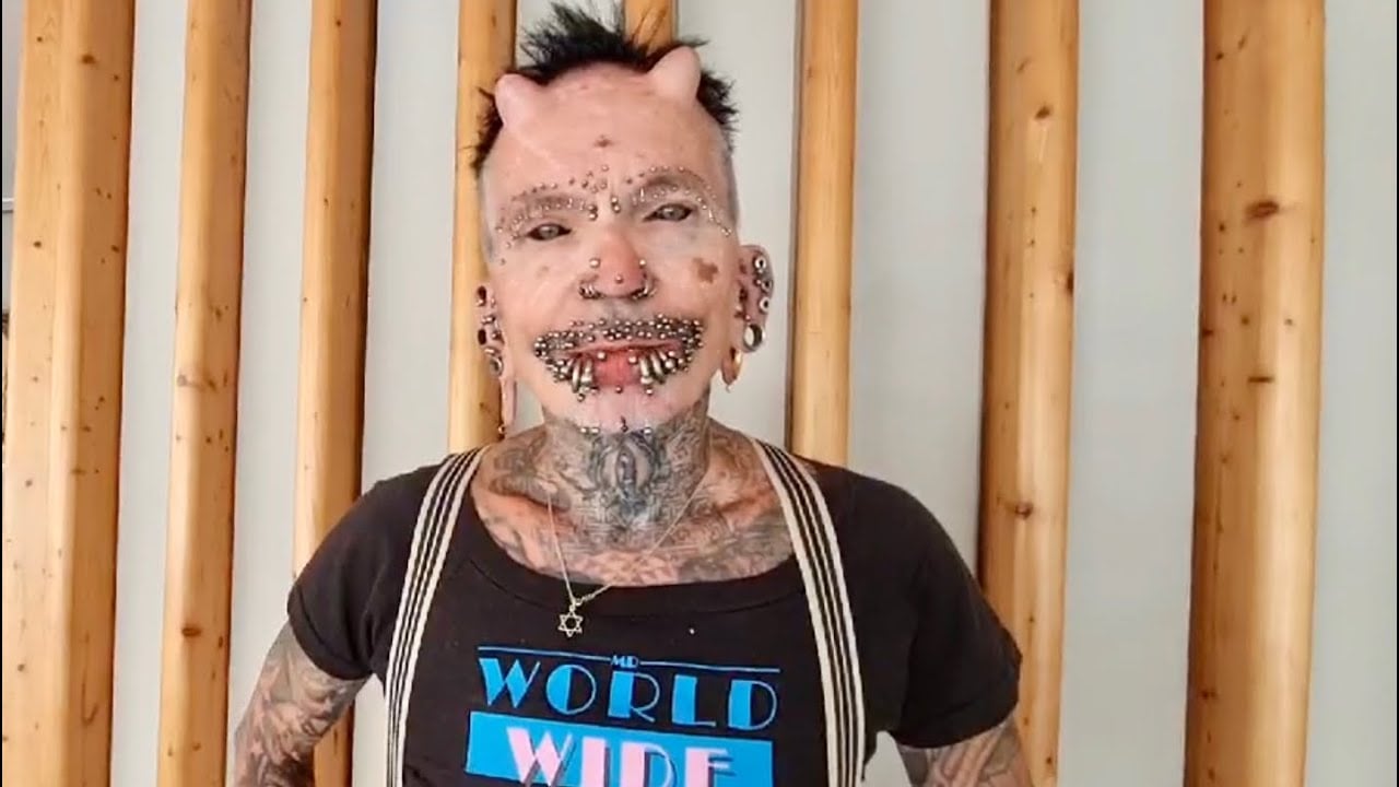 A person with body modifications including facial piercings, subdermal horn implants, and tattoos stands in front of vertical wooden beams, wearing a "WORLD WIDE" t-shirt and suspenders.