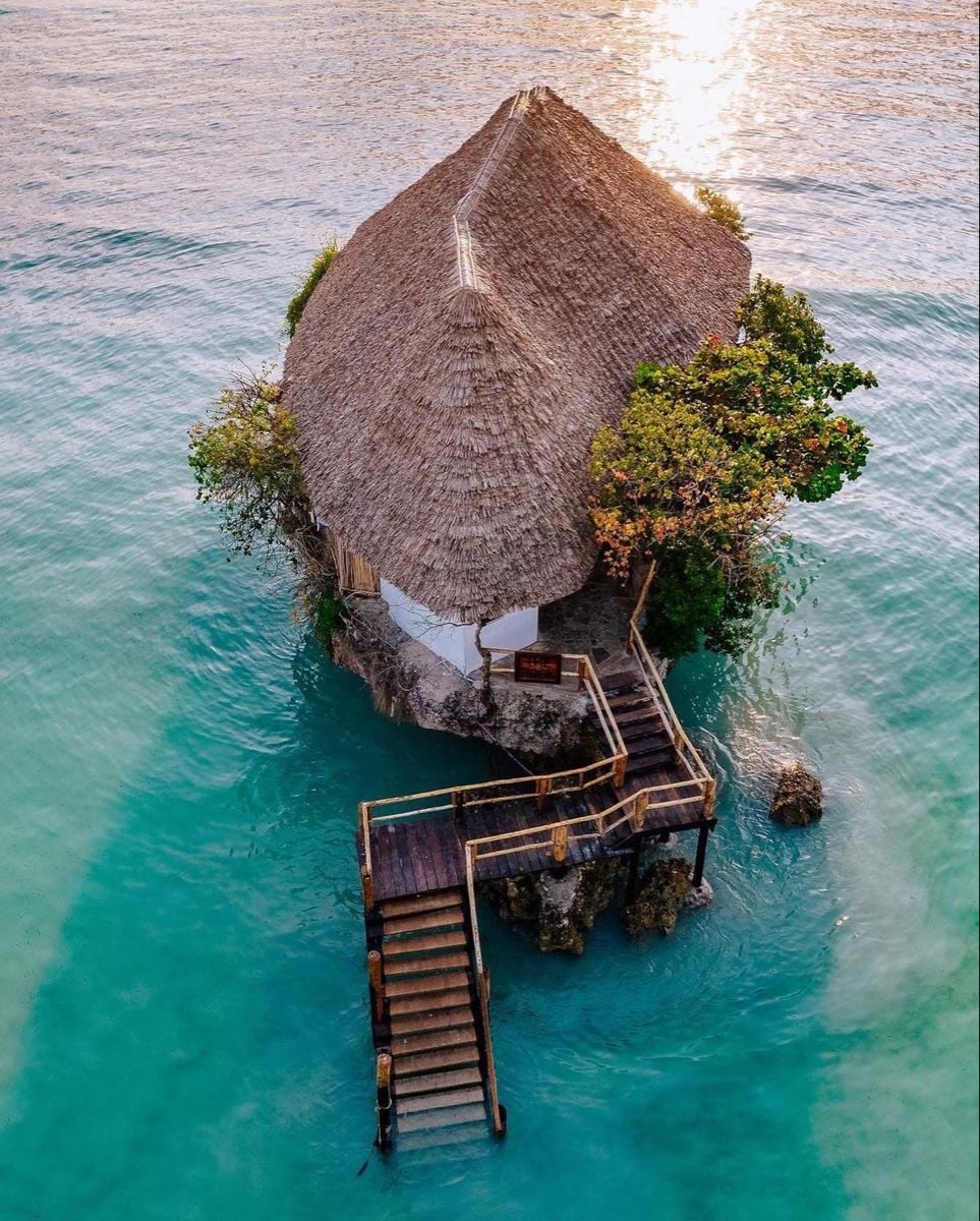 A thatched-roof building sits on a small rock island in turquoise water, connected to the shore by a wooden staircase and deck, surrounded by lush greenery and bathed in sunlight.