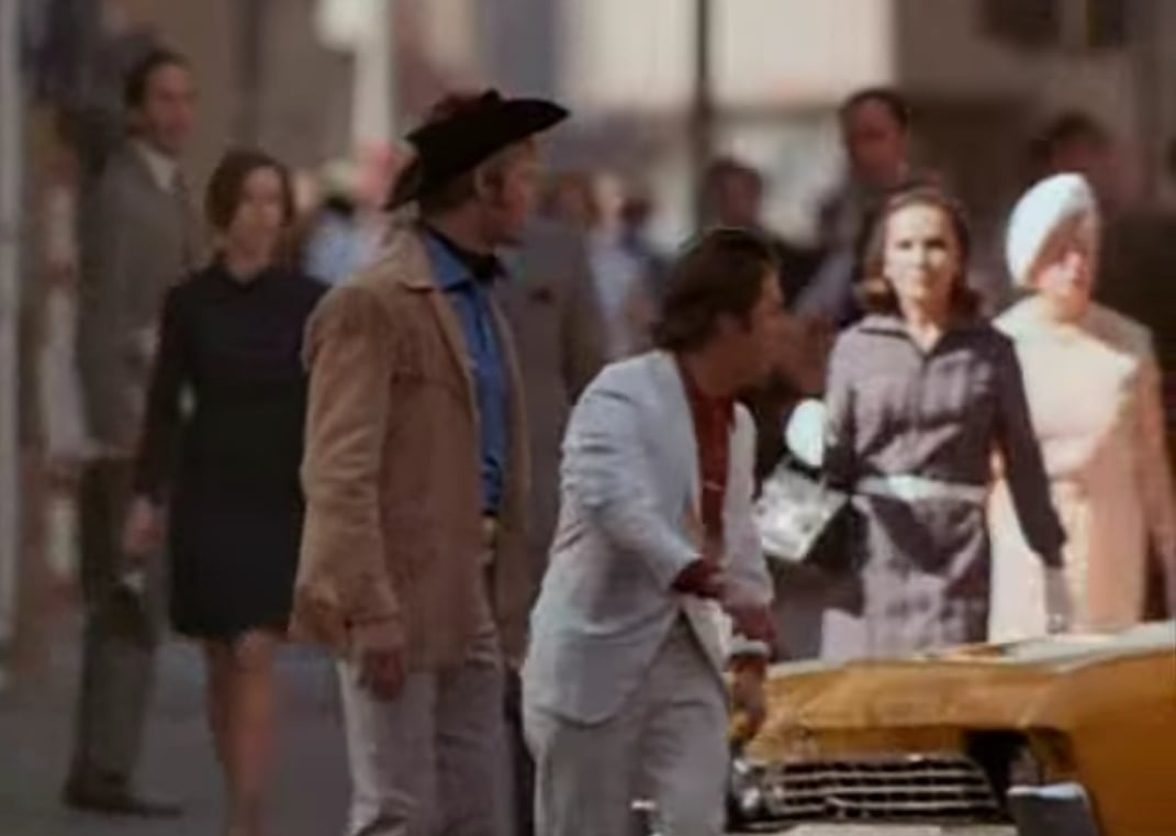Two men cross a busy city street; one wears a cowboy hat and tan jacket, the other wears a white suit. Pedestrians in 1960s-style clothing walk on the sidewalk in the background, and a yellow taxi is partially visible.