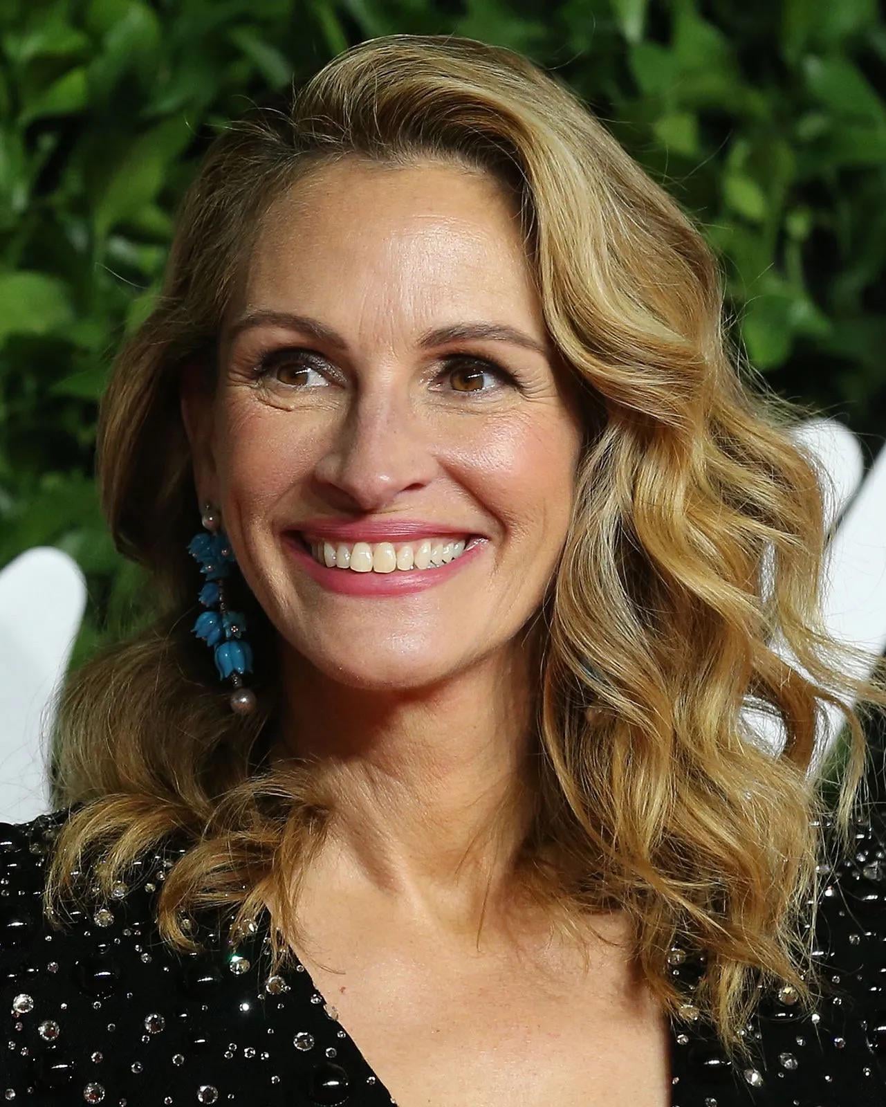 A woman with wavy blonde hair, wearing a black sequined top and blue statement earrings, smiles in front of a leafy green background.