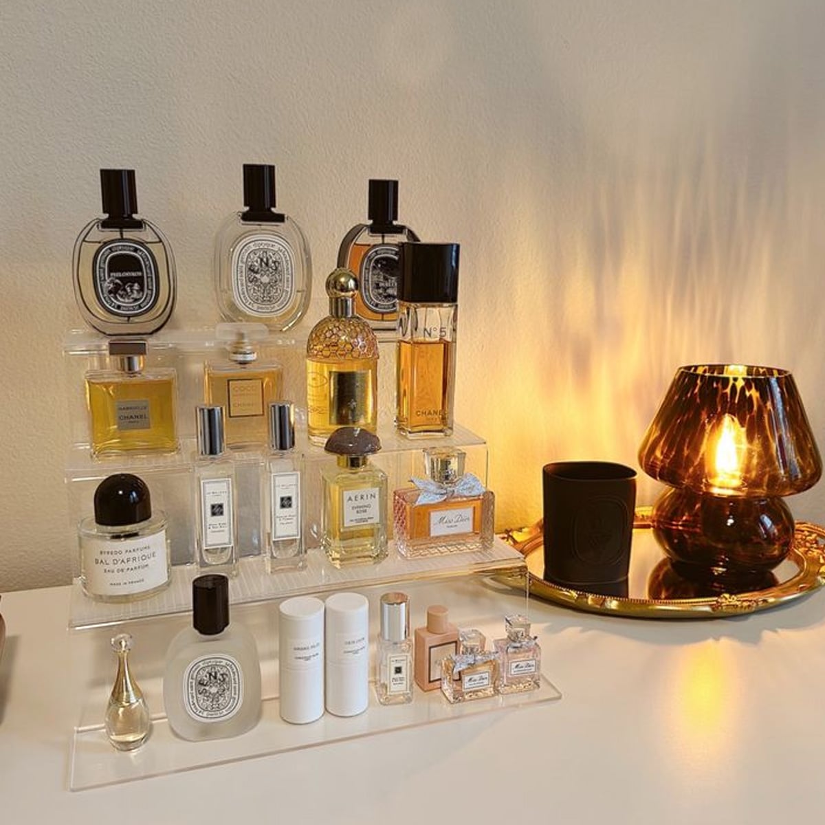 A collection of perfumes arranged on a clear acrylic organizer, displayed on a white surface next to a glowing amber glass lamp and a black candle on a gold tray. The setup gives a cozy and elegant vibe.