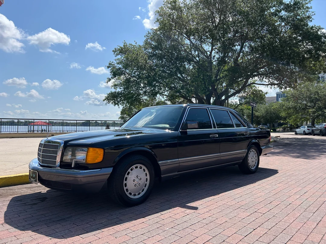 A classic dark-colored Mercedes-Benz sedan is parked on a brick road in the shade of a large tree, with a sunny sky and riverfront railing visible in the background.