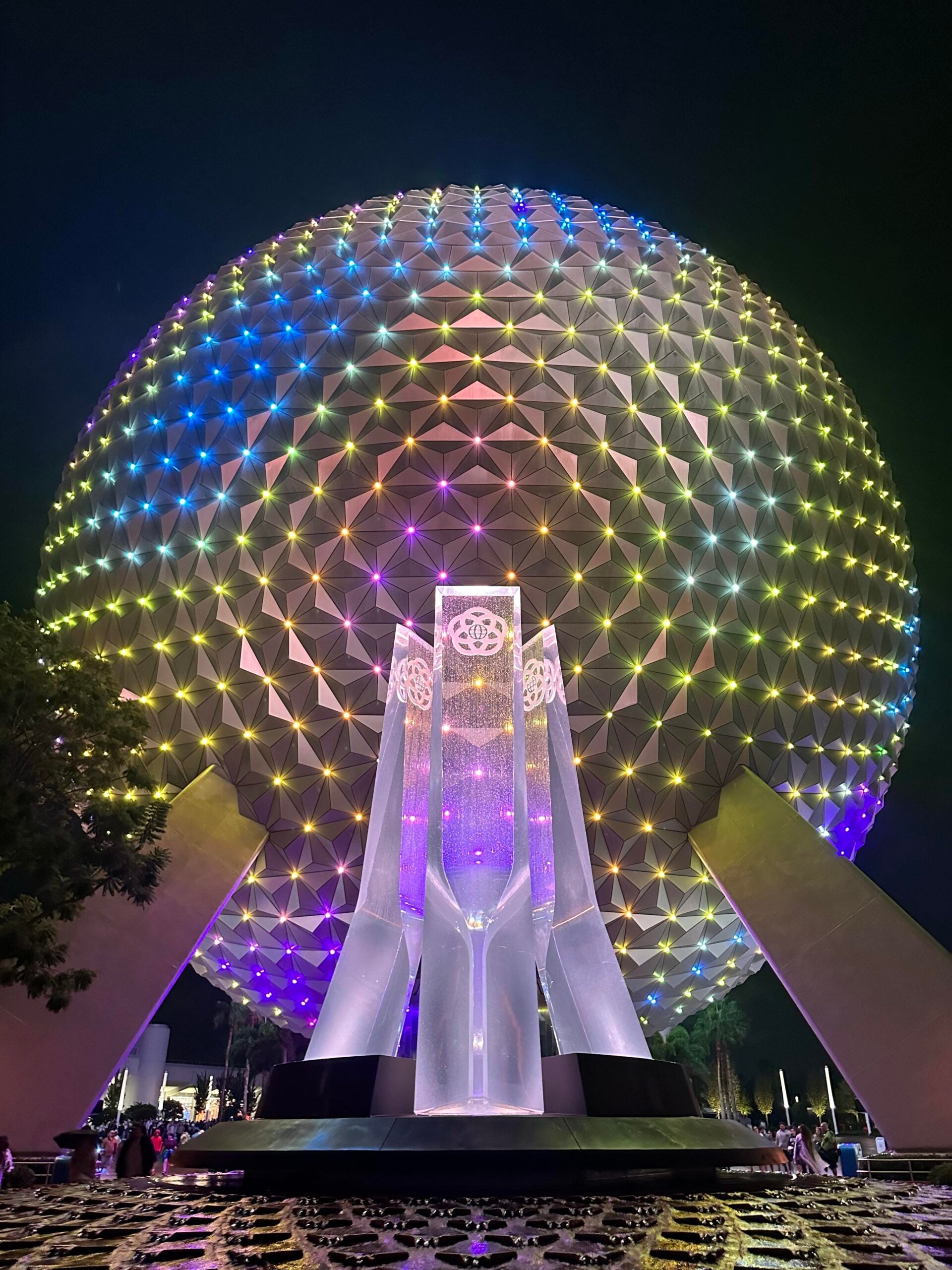 A large geodesic sphere, Spaceship Earth at EPCOT, glows with colorful lights at night. In front of it is a tall, illuminated glass monument with patterns and symbols, and a water fountain at the base.