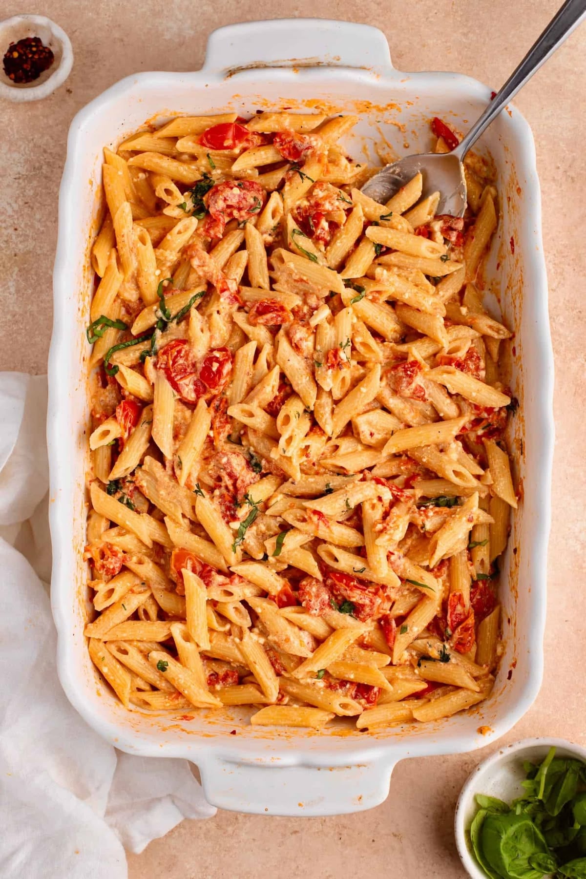 A white baking dish filled with creamy baked penne pasta, cherry tomatoes, spinach, and herbs, with a serving spoon resting inside. Small bowls of red pepper flakes and fresh basil sit nearby.
