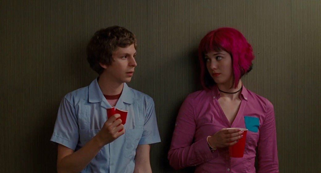 Two young people stand against a plain wall, each holding a red cup. The person on the left wears a light blue shirt, while the person on the right has bright pink hair and a pink shirt. They are looking at each other.