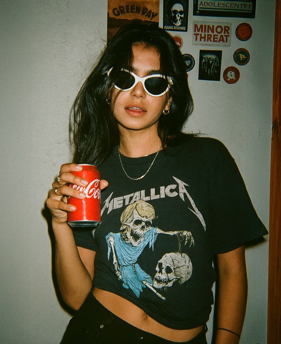 A woman wearing white sunglasses and a black Metallica t-shirt holds a can of Coca-Cola. Behind her, a wall is decorated with band stickers and pins, including “MINOR THREAT” and “ADOLESCENTS.”