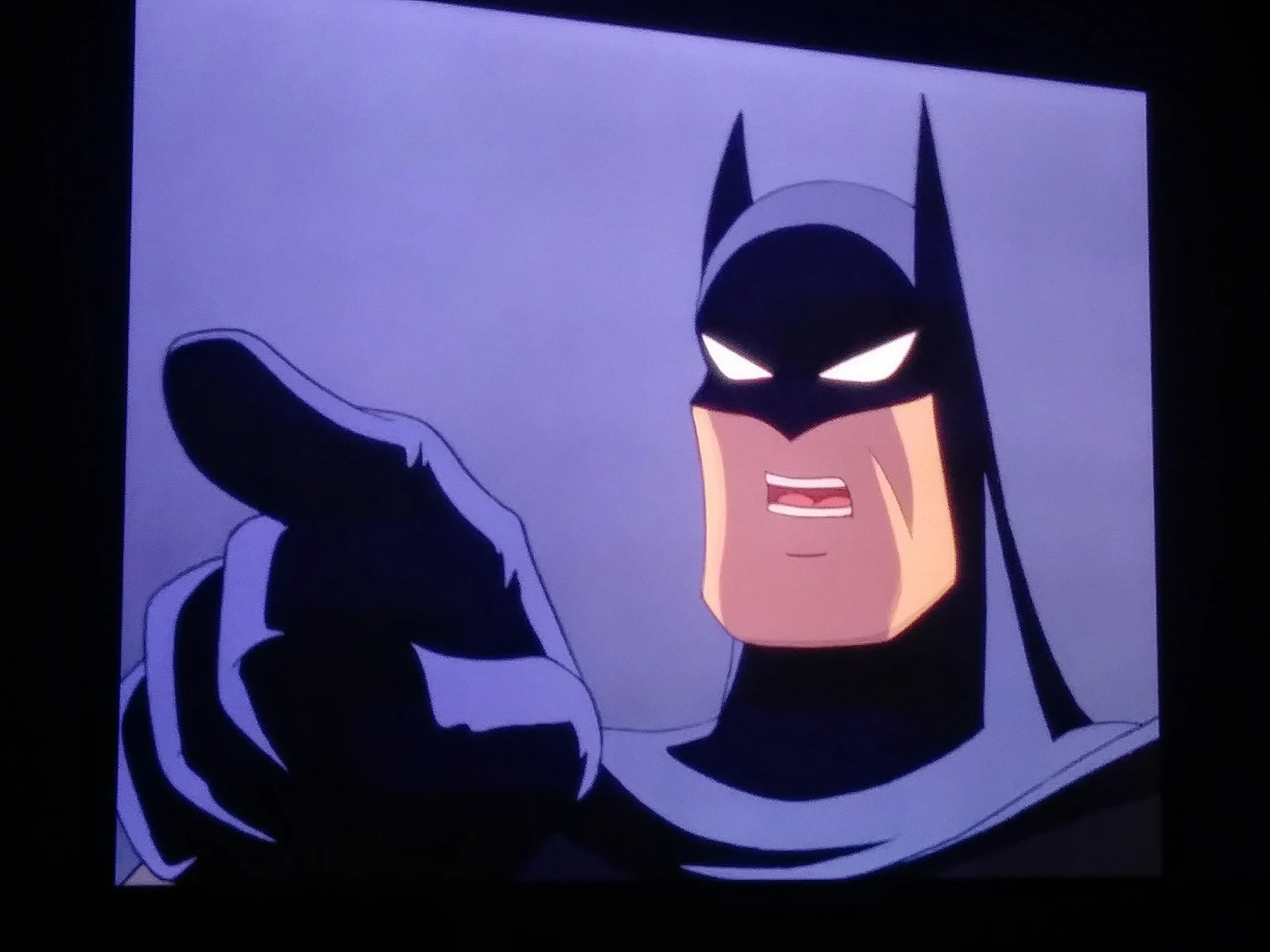 Animated Batman in dark costume is pointing forward with a stern expression on his face against a plain, purple background.