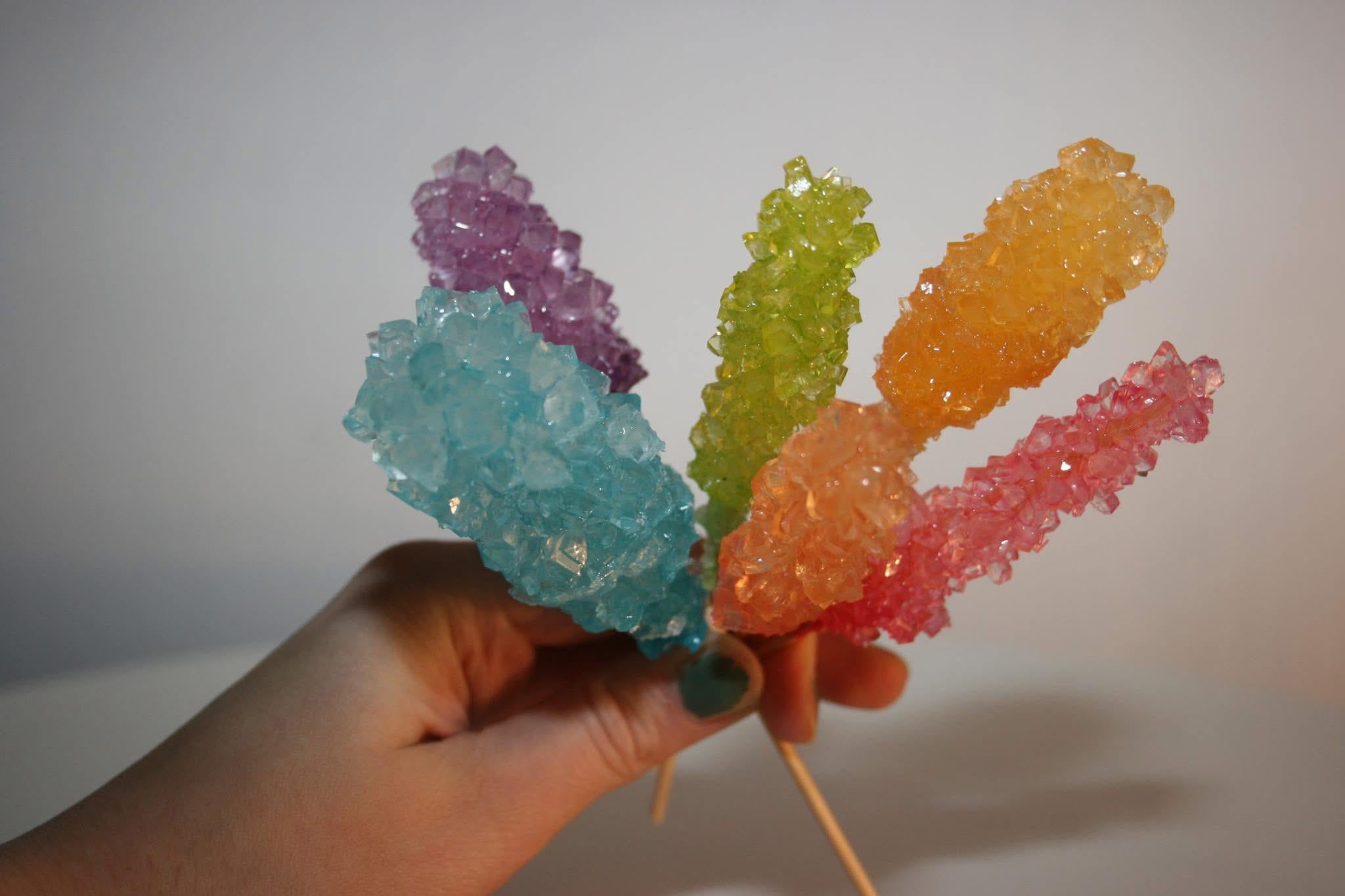 A hand holding five colorful rock candy sticks, each with large sugar crystals in blue, purple, green, orange, and pink, against a plain light background.