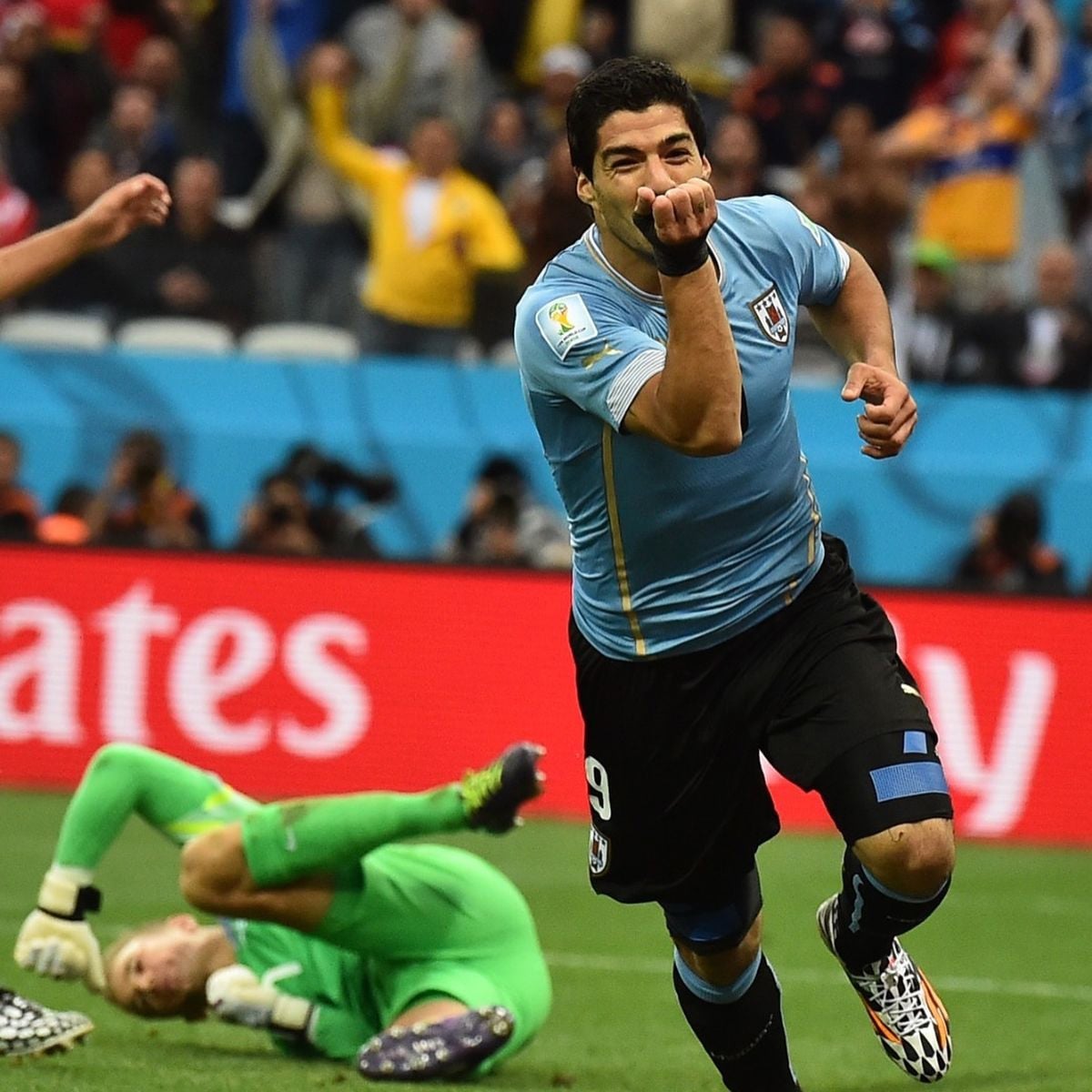 A soccer player in a light blue jersey celebrates after scoring a goal, running with a hand gesture towards his mouth, while a goalkeeper in green lies on the ground in the background.