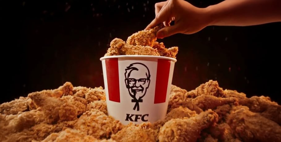 A hand reaches into a large KFC bucket filled with crispy fried chicken, surrounded by more pieces of fried chicken, against a dark background.