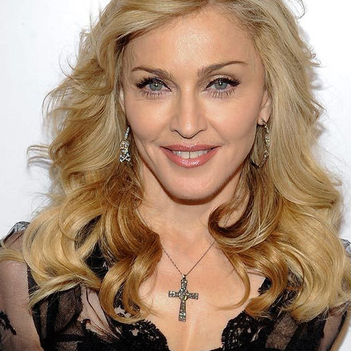 A woman with long, wavy blonde hair wears a black lace top, large hoop earrings, and a cross necklace, smiling softly at the camera against a plain background.
