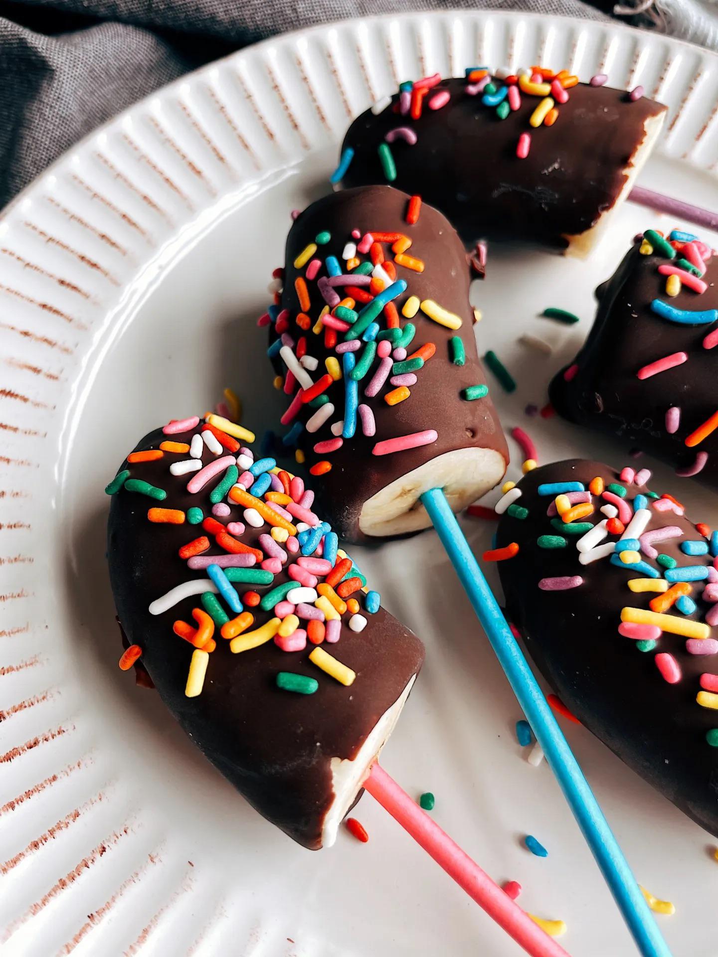 Chocolate-covered banana slices on sticks, topped with colorful rainbow sprinkles, arranged on a white plate.