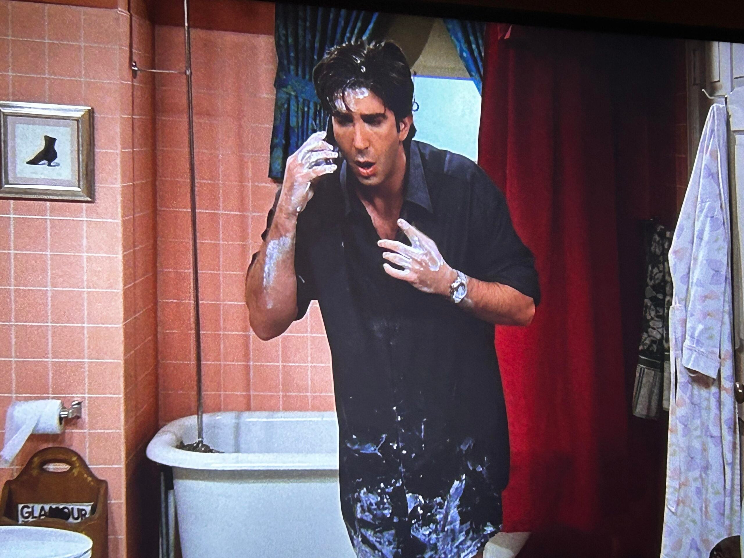 A man standing in a bathroom is covered in white powder, looking surprised and confused, with powder on his hands, shirt, and pants. A clawfoot bathtub and pink tiled walls are visible in the background.