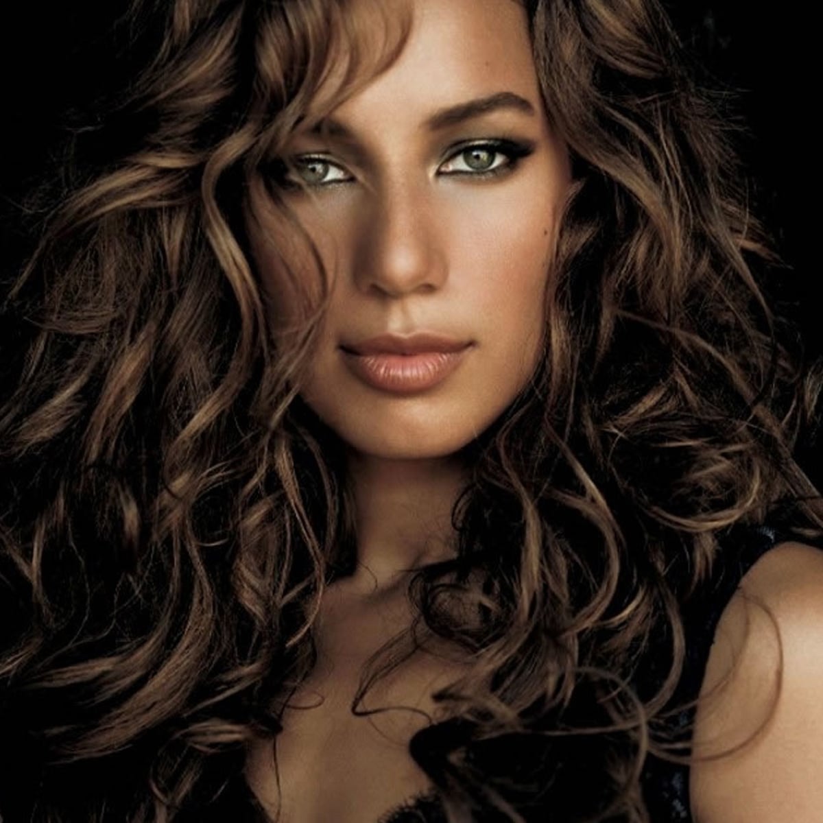 A woman with long, voluminous curly brown hair and light eyes gazes directly at the camera. She has soft makeup and is shown against a dark background.