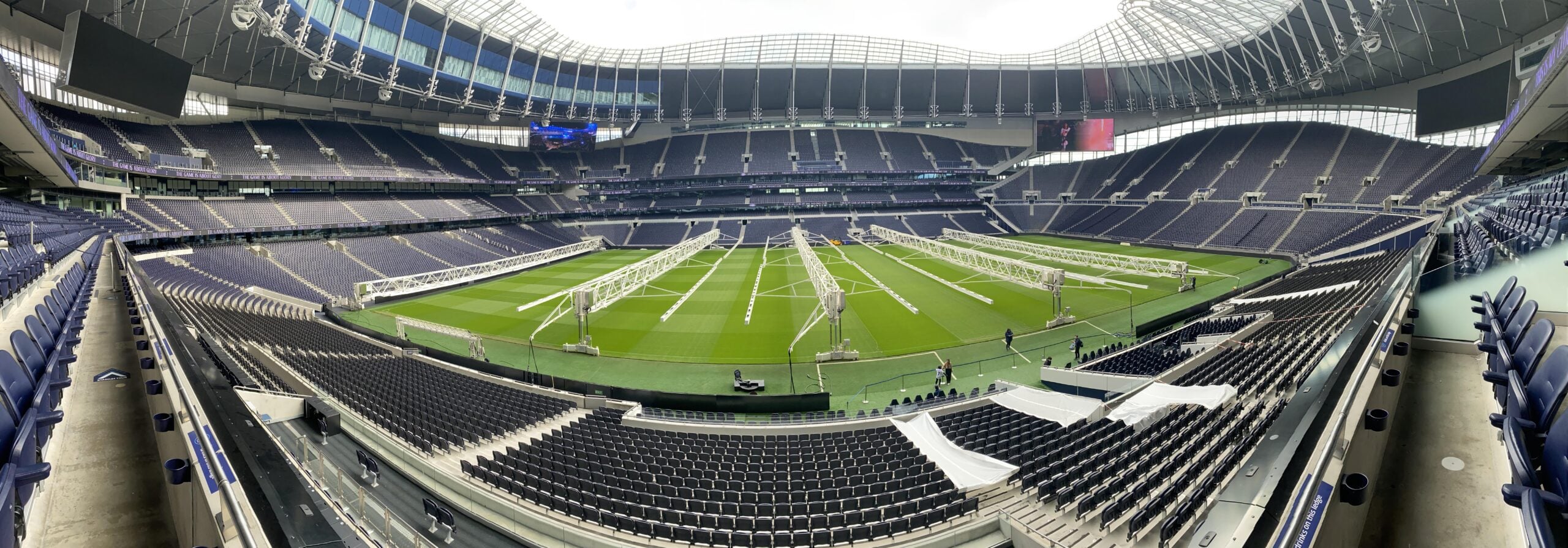 Wide-angle view of an empty, modern football stadium with a green pitch, white grow lights on the field, and rows of dark seats surrounding the playing area. The retractable roof structure is visible above.