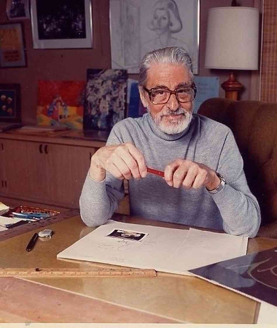 An older man with gray hair and glasses, wearing a light gray turtleneck, sits at a desk holding a red pencil. Art supplies and drawings are on the desk, and colorful artwork decorates the background.