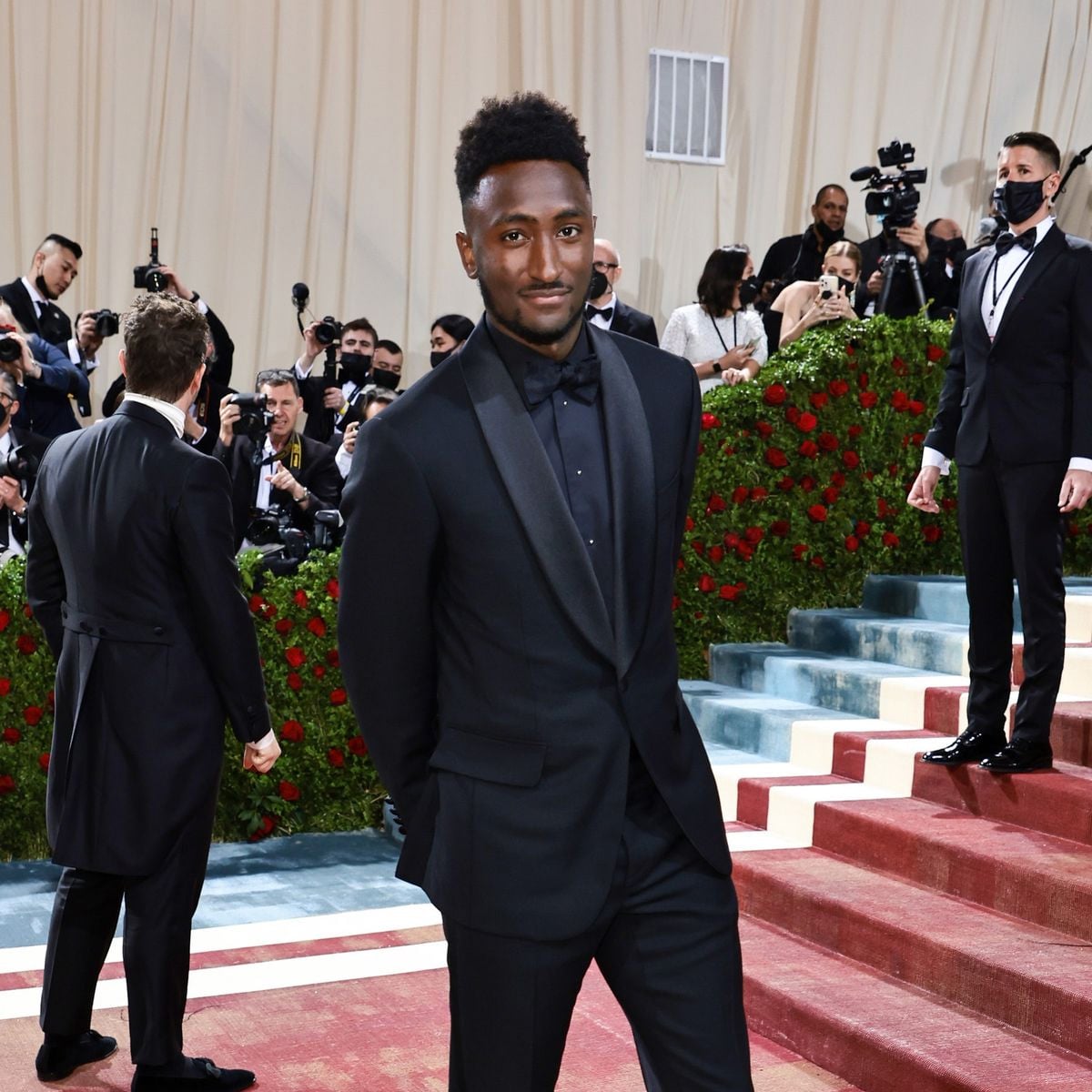 A man in a black tuxedo poses with his hands in his pockets on a red carpeted staircase, surrounded by photographers and guests at a formal event with red and green floral decorations.