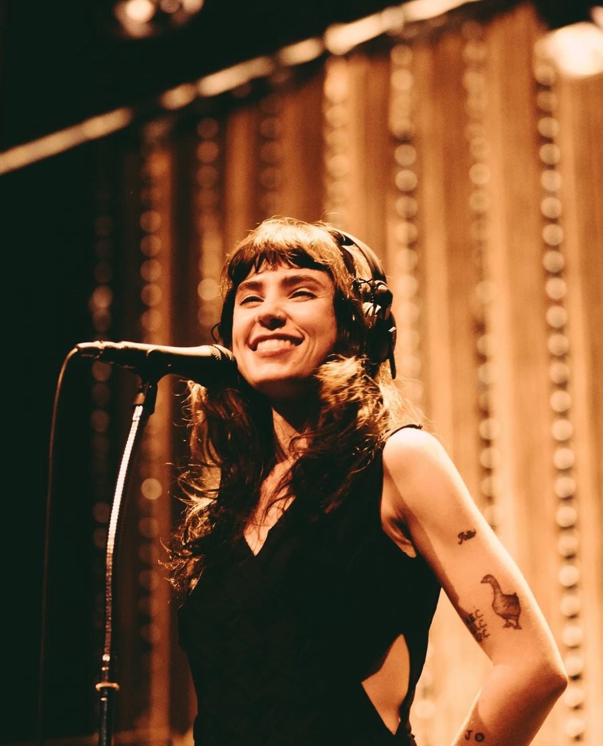 A smiling woman with long dark hair and bangs stands at a microphone, wearing headphones. She has tattoos on her arm and is in front of a warm, softly lit background with vertical light strands.