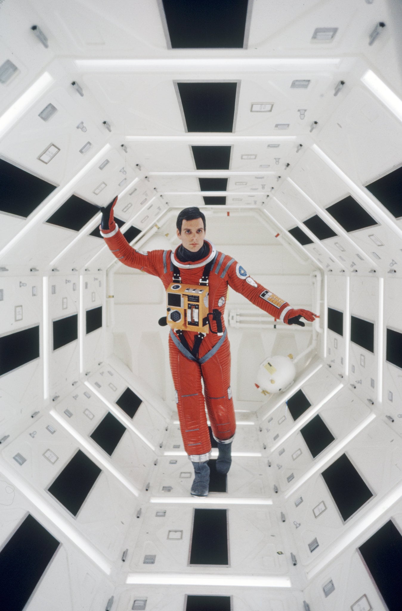An astronaut in a red spacesuit floats inside a white, hexagonal space station corridor with black and light panels on the walls and a helmet by his side.