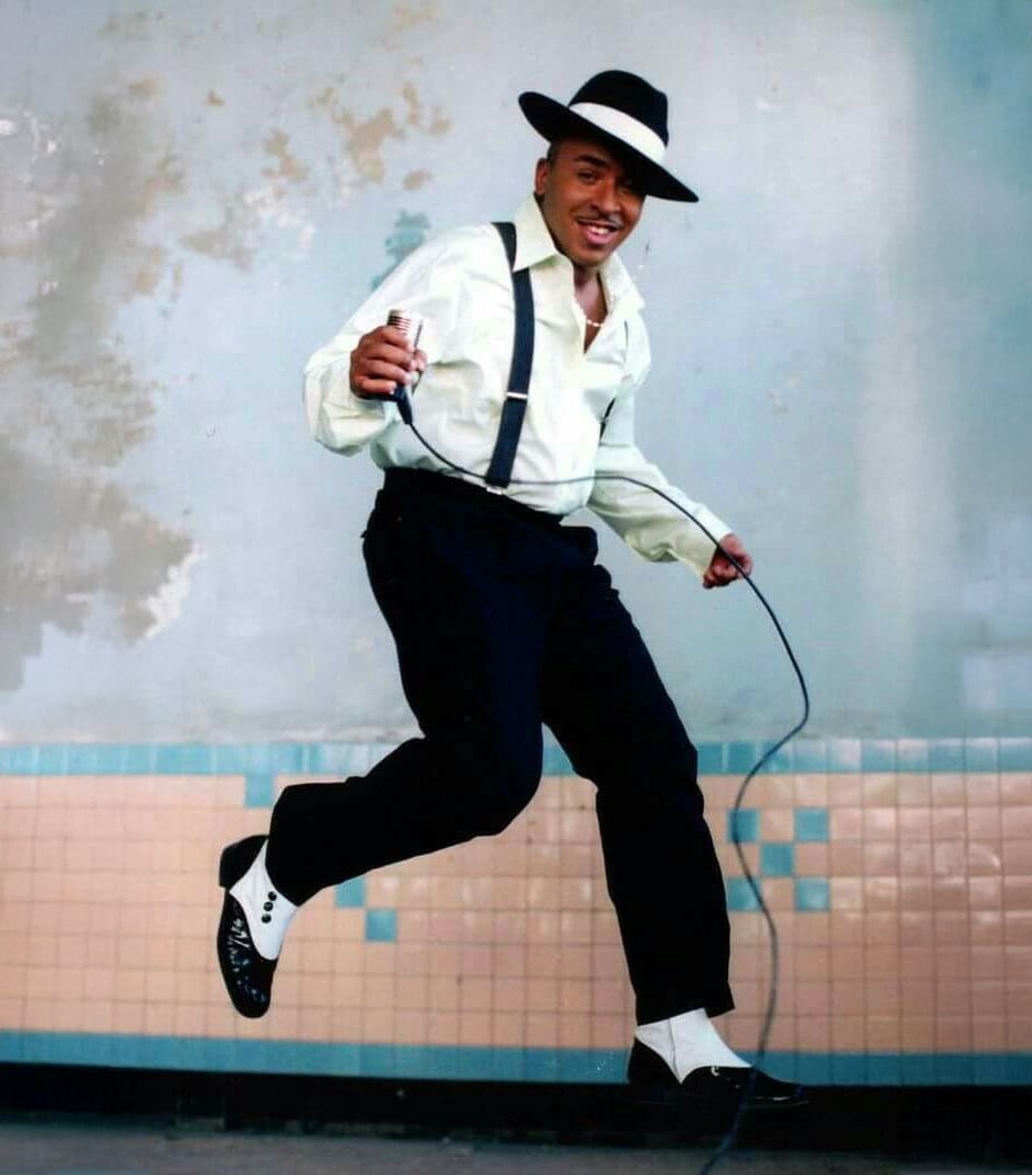 A man dressed in a white shirt, black suspenders, black pants, black-and-white shoes, and a black fedora hat dances joyfully while holding a microphone, against a blue and beige tiled wall.